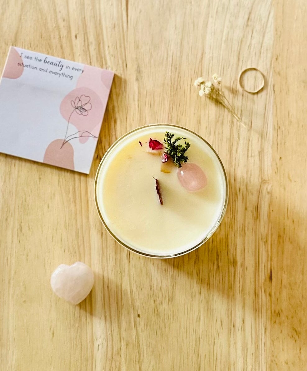 Top view of Love Rose Quartz Crystal Candle with delicate ring, affirmation note pad and rose quartz heart crystal surrounding.