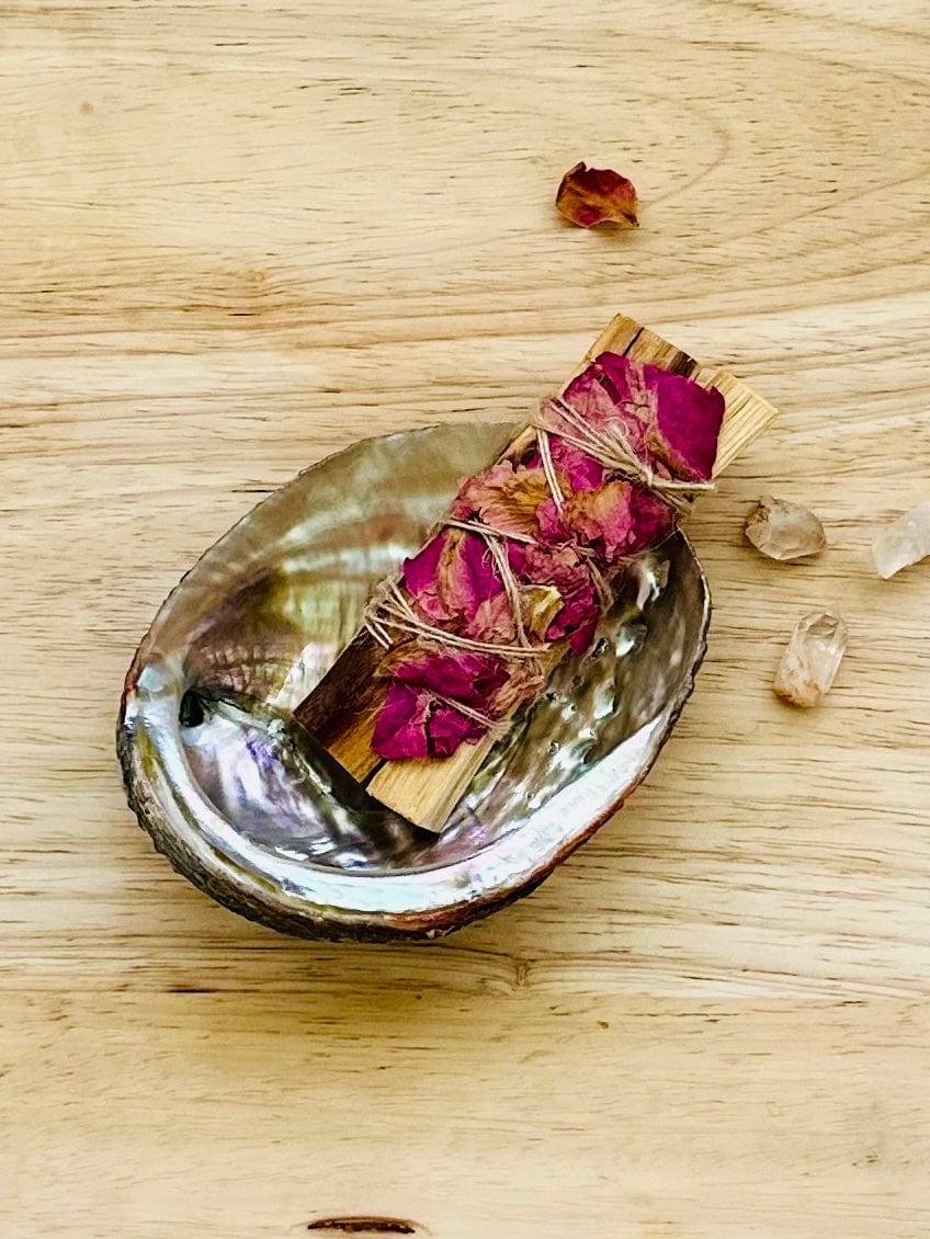 Abalone shell cradling rose-wrapped Palo Santo bundle with crystals.