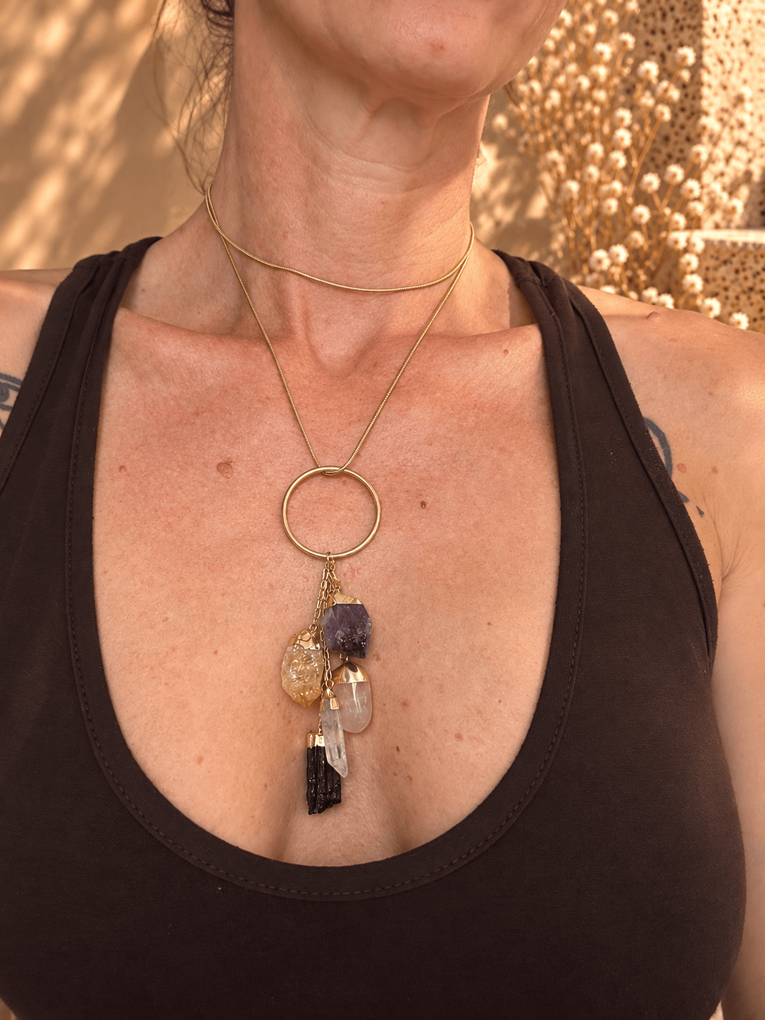 A close-up of a woman wearing a layered necklace with a gold-tone ring and dangling crystal charms, including amethyst and black tourmaline, over a dark brown tank top.
