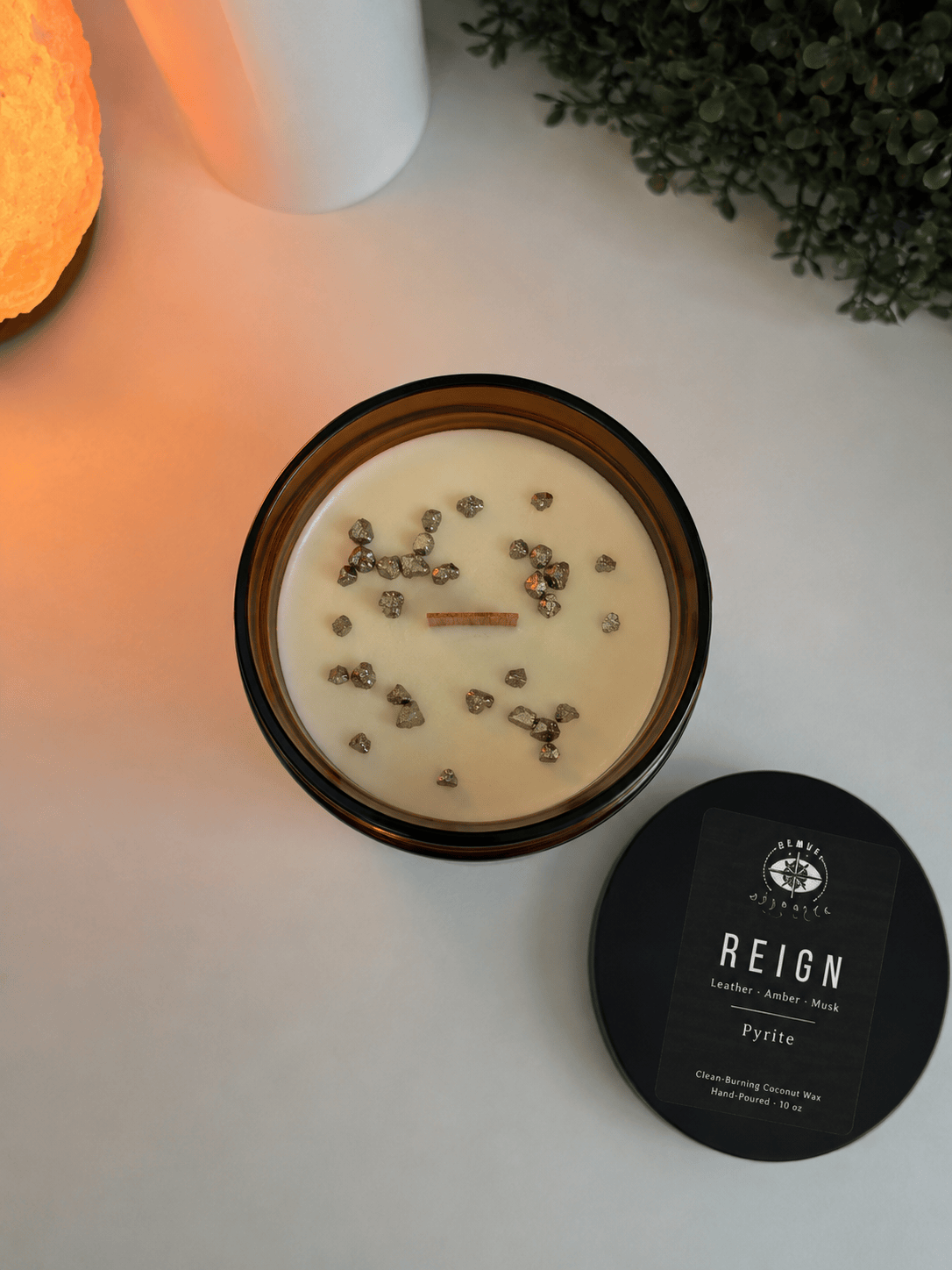 Candle in a black container with visible herbs on a white surface, next to a black lid labeled &
