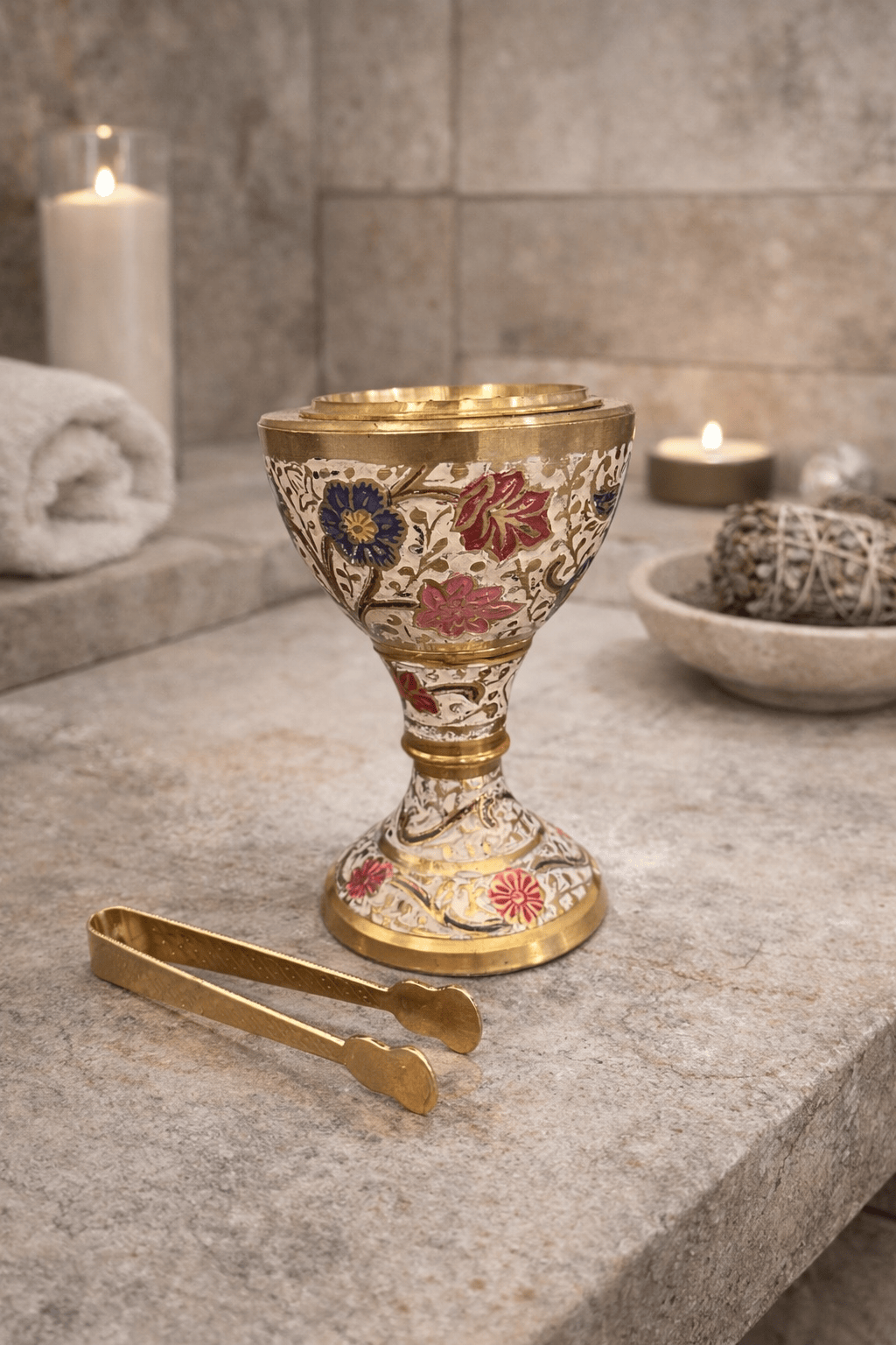 Floral brass incense burner with enamel design and charcoal tongs set on a ritual altar surface with candles and sage.