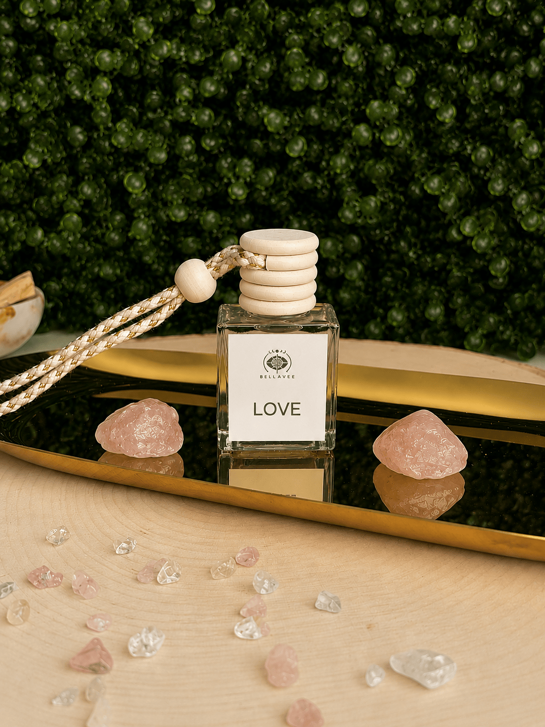 BellaVee LOVE crystal car diffuser displayed on a gold mirrored tray with two polished rose quartz crystals. The square glass diffuser features a natural wood cap and braided hanging cord, set against a lush green botanical backdrop with scattered rose quartz and clear quartz crystal chips. A romantic fragrance diffuser designed to bring warmth, self-love, and calming energy to any vehicle.