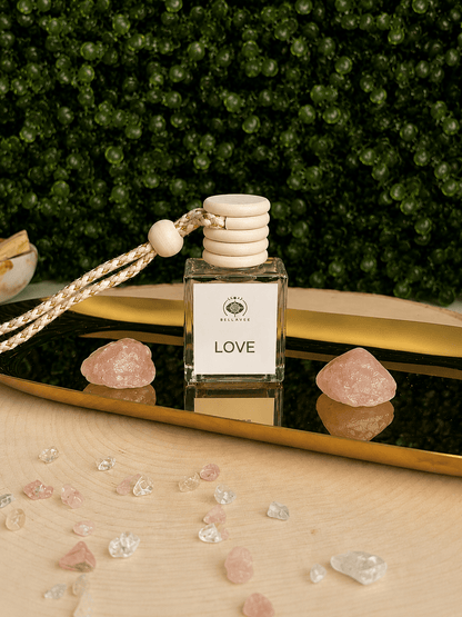 BellaVee LOVE crystal car diffuser displayed on a gold mirrored tray with two polished rose quartz crystals. The square glass diffuser features a natural wood cap and braided hanging cord, set against a lush green botanical backdrop with scattered rose quartz and clear quartz crystal chips. A romantic fragrance diffuser designed to bring warmth, self-love, and calming energy to any vehicle.