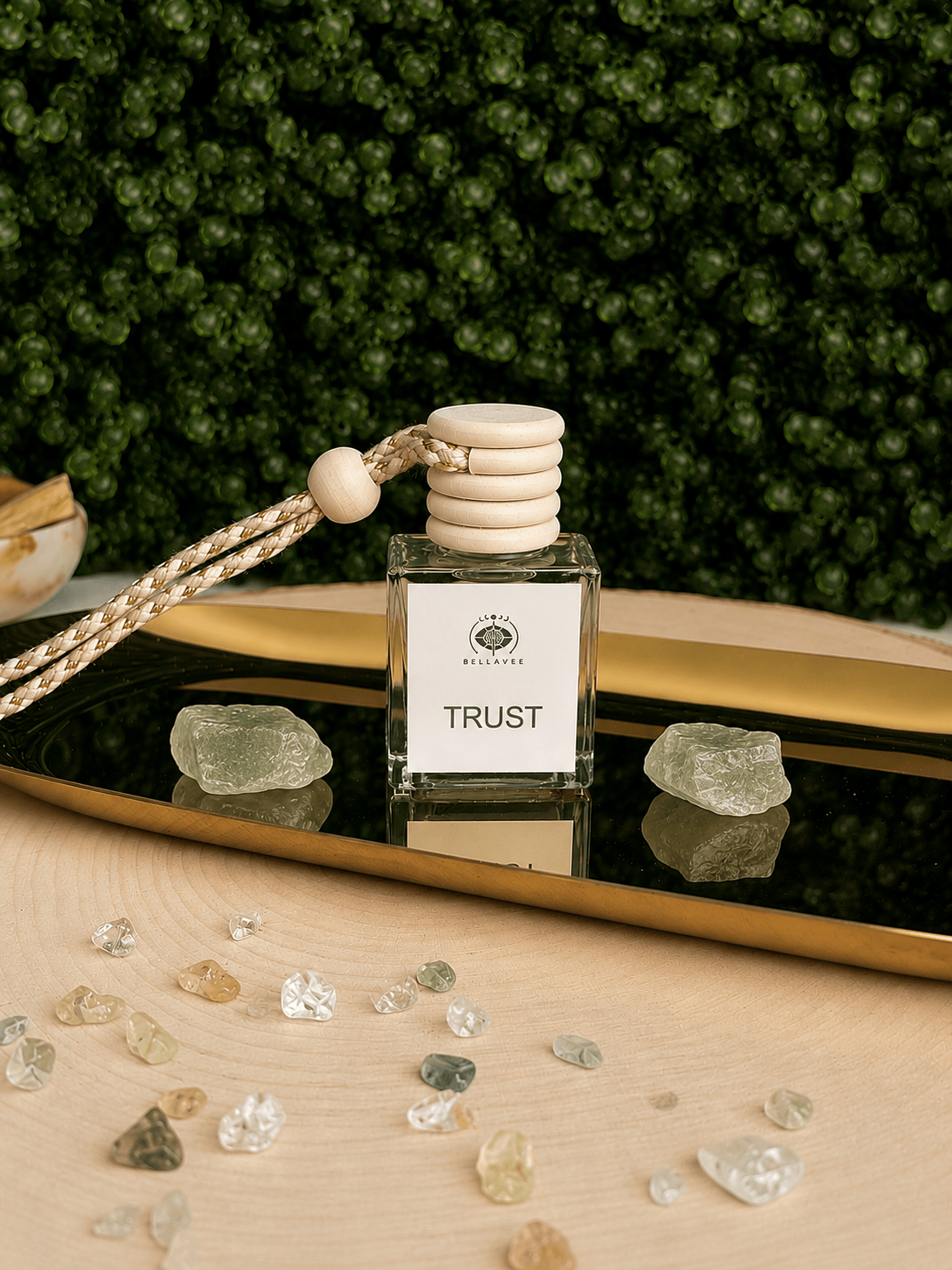 BellaVee Trust crystal car diffuser displayed on a reflective gold tray with two pale green crystals, scattered crystal chips, and a lush green botanical backdrop. The square glass diffuser features a natural wooden cap and braided hanging cord, creating a calming, elegant wellness aesthetic.
