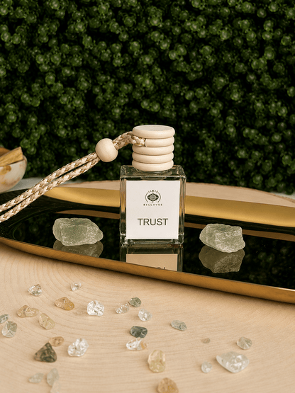 BellaVee Trust crystal car diffuser displayed on a reflective gold tray with two pale green crystals, scattered crystal chips, and a lush green botanical backdrop. The square glass diffuser features a natural wooden cap and braided hanging cord, creating a calming, elegant wellness aesthetic.
