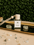 BellaVee Trust crystal car diffuser displayed on a reflective gold tray with two pale green crystals, scattered crystal chips, and a lush green botanical backdrop. The square glass diffuser features a natural wooden cap and braided hanging cord, creating a calming, elegant wellness aesthetic.
