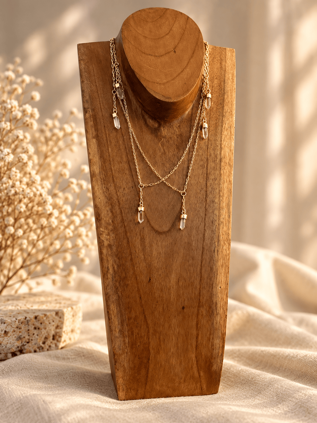 A gold-toned layered necklace with clear teardrop crystal pendants displayed on a wooden neck bust, set against a softly lit neutral fabric background with dried flowers.