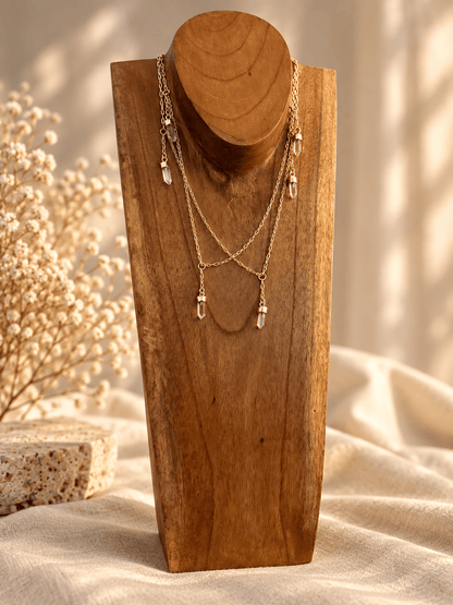 A gold-toned layered necklace with clear teardrop crystal pendants displayed on a wooden neck bust, set against a softly lit neutral fabric background with dried flowers.