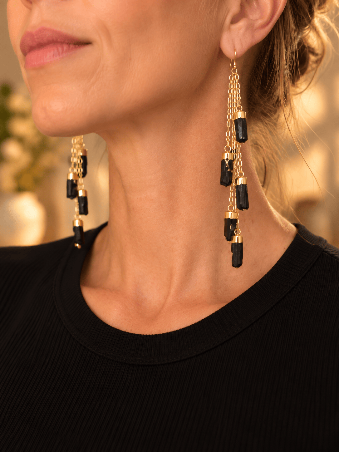 Close-up of a woman wearing long dangling earrings with black tourmaline crystal and gold chain links, paired with a black ribbed top.