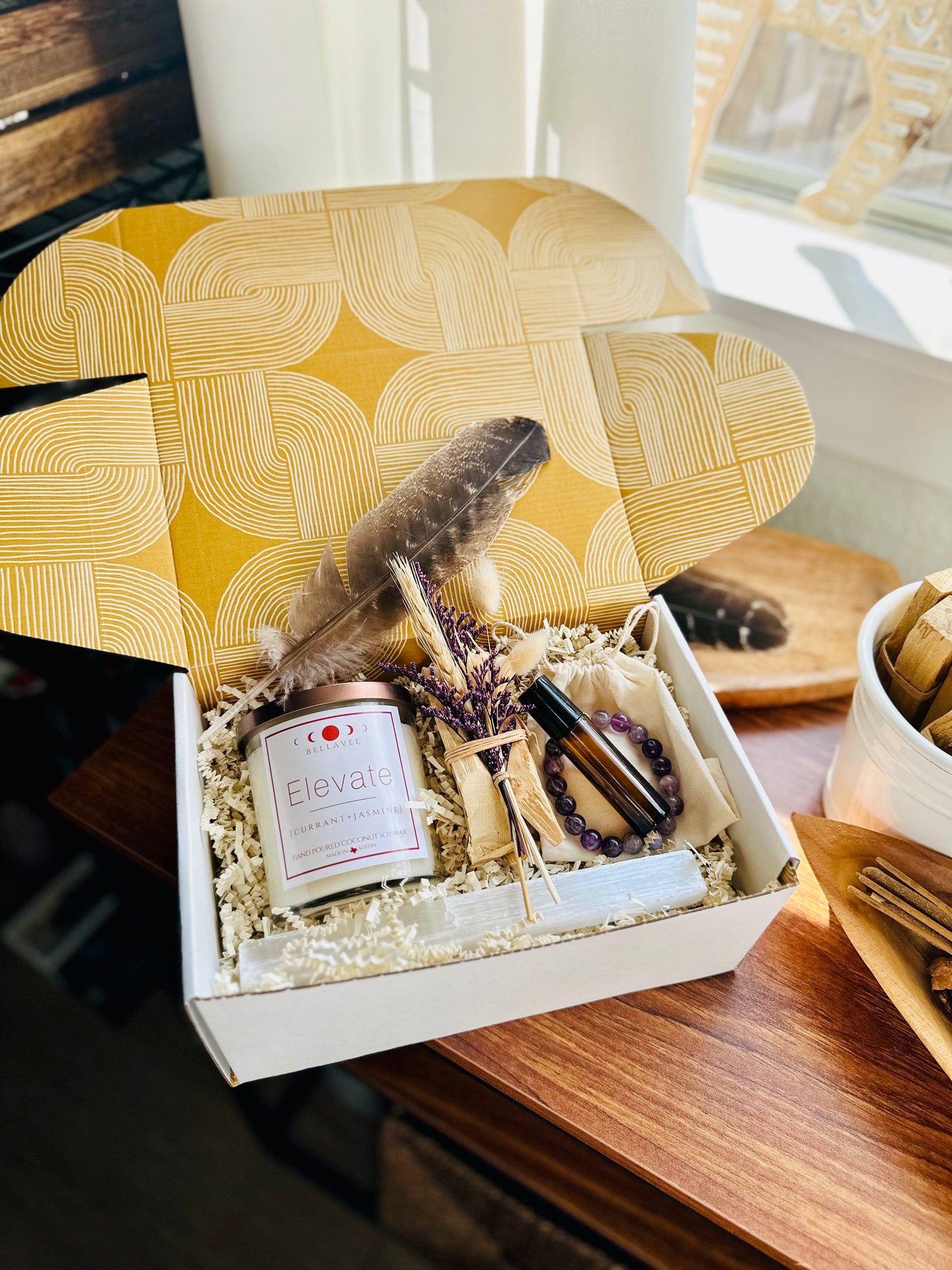 Elevate Amethyst topped crystal Candle wellness gift box with essential oil roll on and Palo Santo bundle with amethyst bracelet and selenite wand. sitting on wooden table with window lighting. Purple accents beautifully packaged.