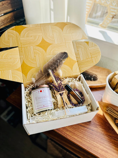 Elevate Amethyst topped crystal Candle wellness gift box with essential oil roll on and Palo Santo bundle with amethyst bracelet and selenite wand. sitting on wooden table with window lighting. Purple accents beautifully packaged.