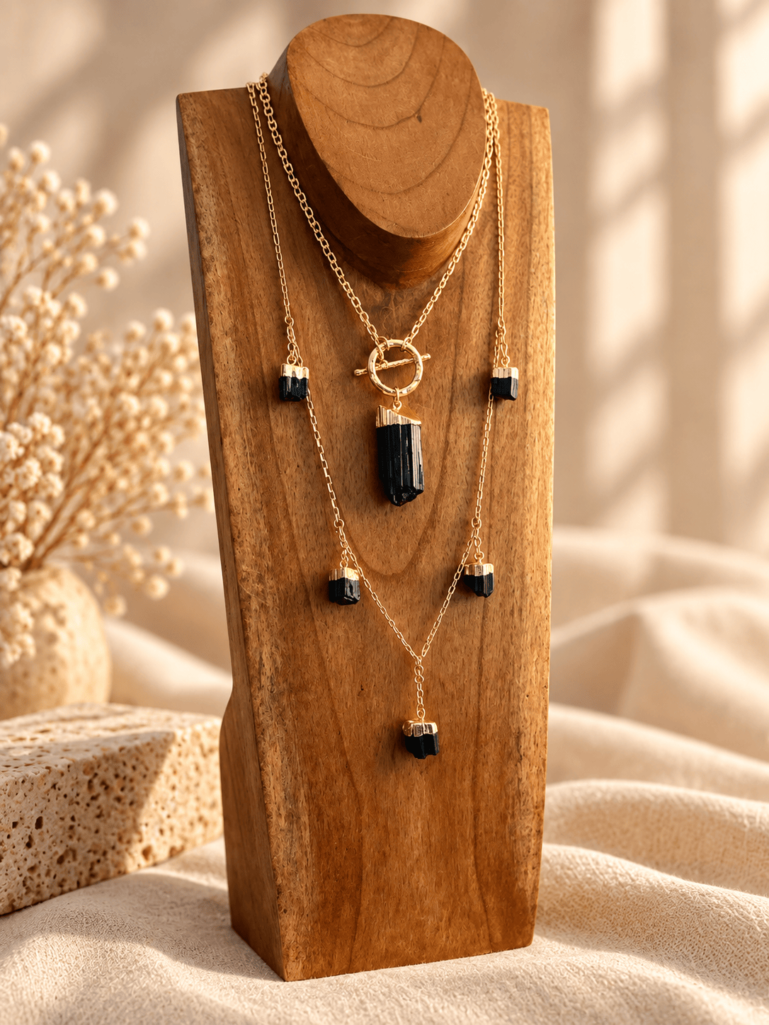 A gold layered necklace set with Black Tourmaline crystal pendant displayed on a wooden stand against a warm beige background with soft shadows.