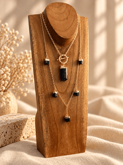 A gold layered necklace set with Black Tourmaline crystal pendant displayed on a wooden stand against a warm beige background with soft shadows.