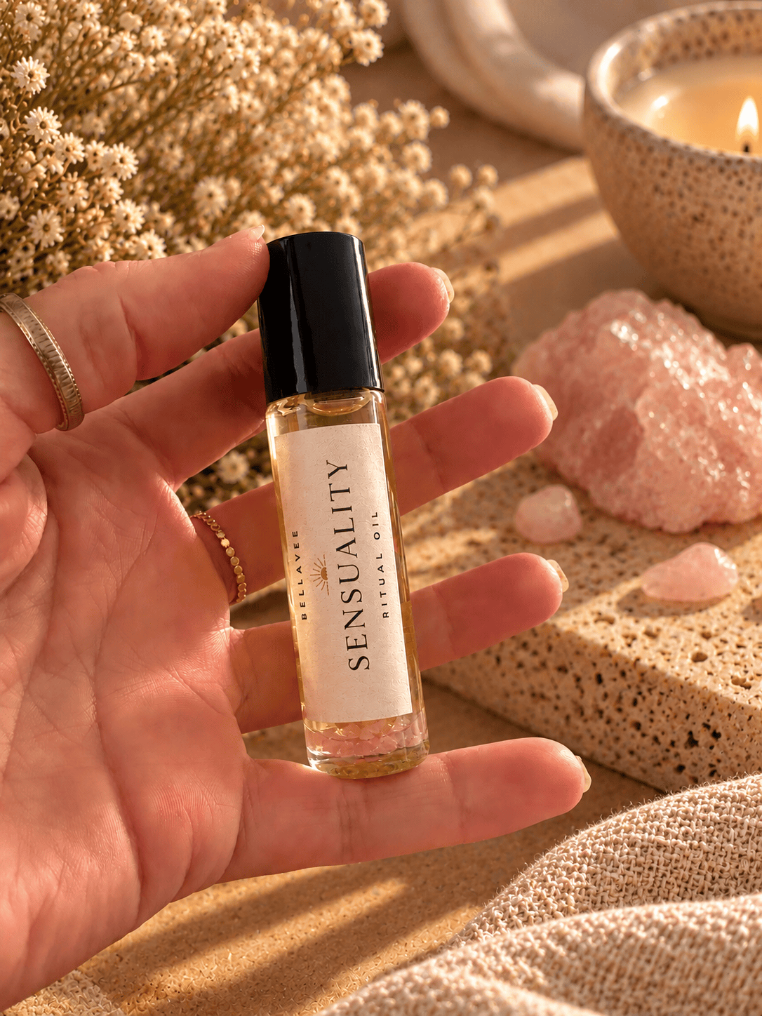 Close-up of a woman holding BellaVee SENSUALITY Ritual Oil roll-on with rose quartz chips against a warm, neutral background with soft natural lighting.