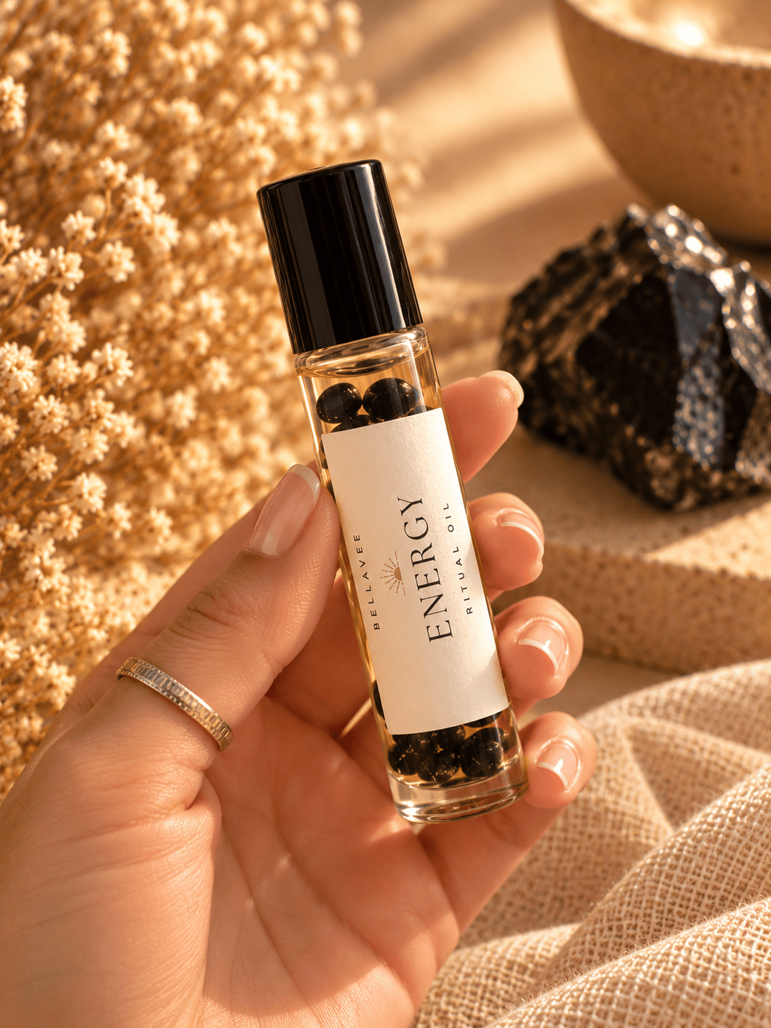 Close-up of a woman holding BellaVee ENERGY Ritual Oil roll-on with black obsidian chips against a warm, neutral background.