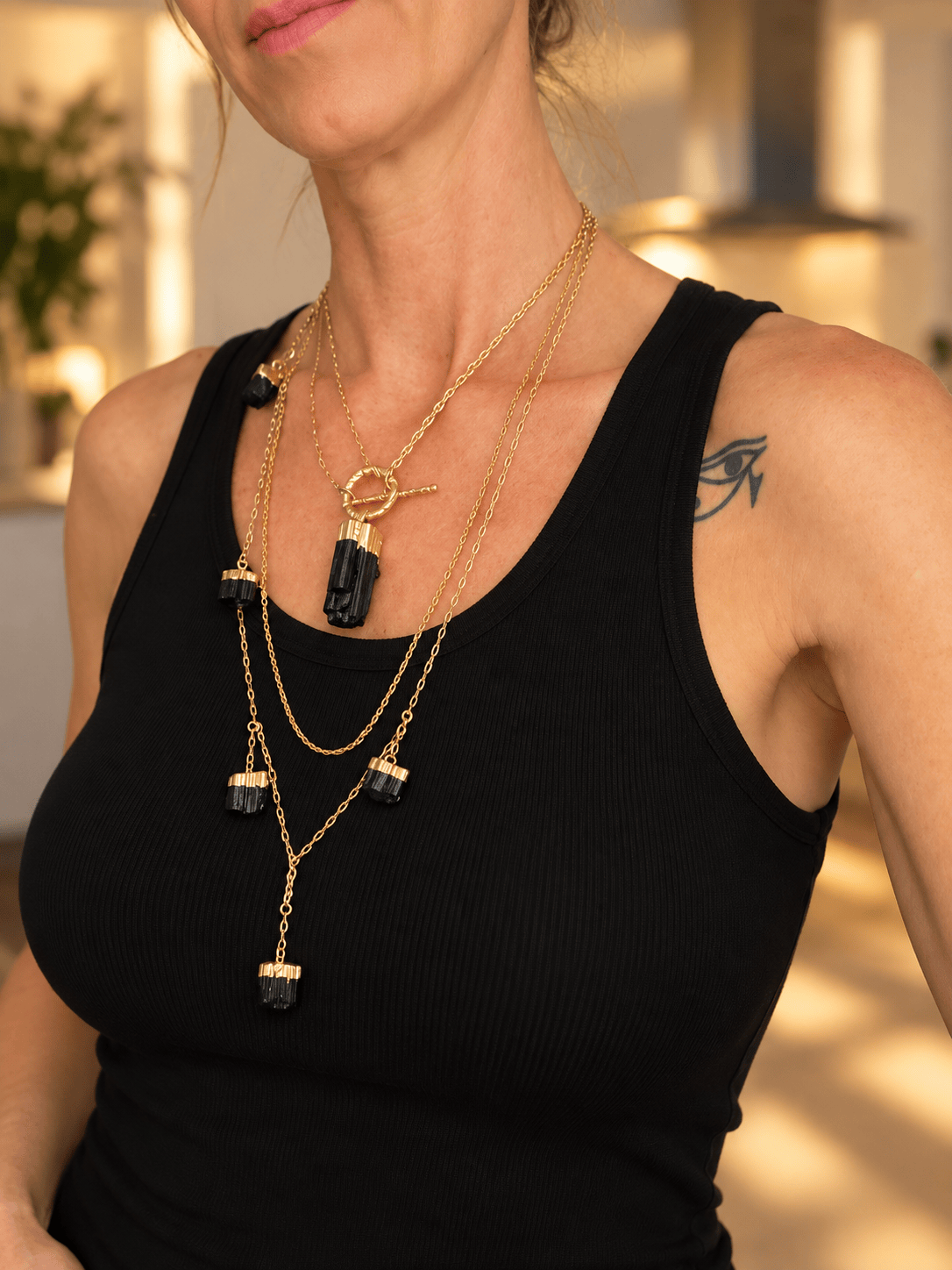 A close-up of a woman wearing a layered necklace set with gold chains and black tourmaline pendant, paired with a black ribbed tank top.