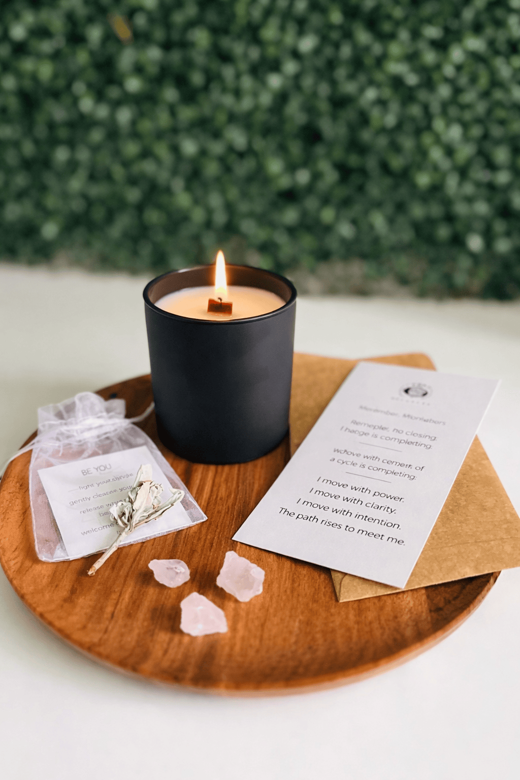 Candle on a wooden tray with rose petals and a card against a green bush background